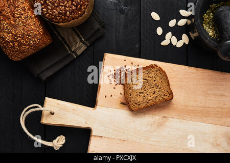 Tagliere con fetta di pane, semi di lino e di vari crostini di pane integrale, semi di zucca e di mortaio su tavoli in legno nero. Vista dall'alto. Foto Stock