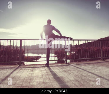 Tall sportivo a riscaldare i suoi muscoli, stirando le sue gambe prima dell'esecuzione di allenamento al mattino. Mattina di sole al di fuori al lago di montagna Foto Stock