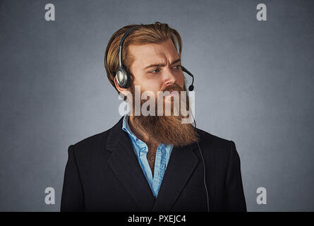 Uomo bello lavorare come un servizio di assistenza clienti, indossa la cuffia alle chiamate, guarda scontroso e annoiato, sconvolto e gravi barbuto hipster business man. Foto Stock