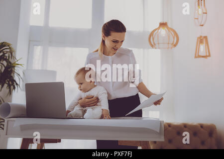 Madre moderna ponendo suo grazioso Baby girl nella parte anteriore del computer portatile mentre si lavora Foto Stock