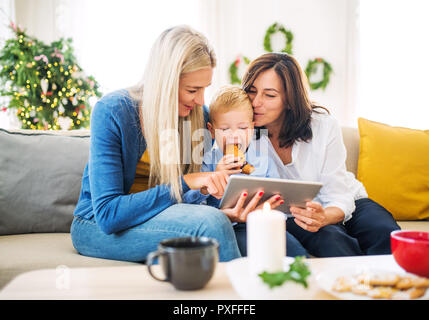 Un piccolo ragazzo con la madre e la nonna seduta su un divano di casa al tempo di Natale, tenendo selfie con la compressa. Foto Stock