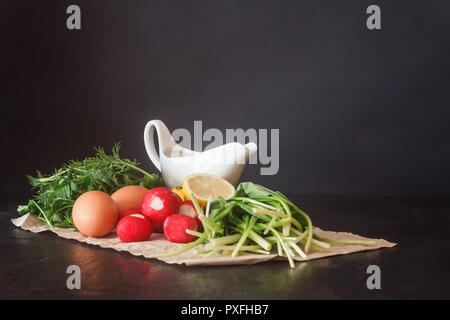 Preparate le verdure fresche e prodotti per insalata, aglio selvatico, Rafano, limone, urkop, uova di gallina, la salsa in un tegame su carta pergamena al buio su un ba Foto Stock