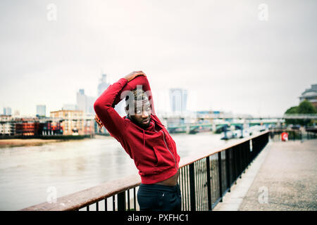 Giovane sportivo da uomo nero runner con smartwatch facendo stretching sul ponte fuori in una città. Foto Stock