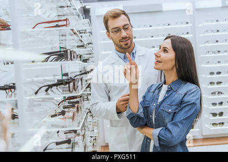 Donna sorridente rivolta dal dito a ripiani con occhiali a maschio all'ottico optometrista in optica Foto Stock