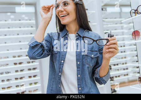 Messa a fuoco selettiva di felice giovane donna scelta di occhiali in negozio oftalmica Foto Stock