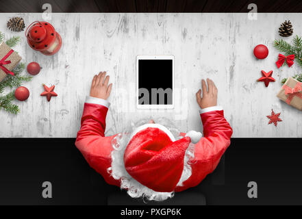 Santa Claus utilizzano tablet con schermo vuoto, per app design presentazione, mockup. Vista scena con decorazioni di Natale accanto a. Foto Stock