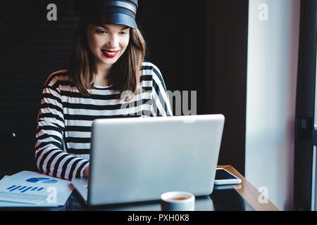 Concentrati sul lavoro. Fiducioso giovane donna in smart casual lavorando sul computer portatile mentre è seduto vicino alla finestra in ufficio creativo o cafe. Foto Stock