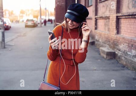 Bella ragazza ridere con lo smartphone dispone di una buona volta in autunno weekend. Outdoor ritratto di amabile alla moda di donna con capelli biondi indossa il cappello in giornata di ottobre Foto Stock