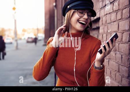 Bella ragazza ridere con lo smartphone dispone di una buona volta in autunno weekend. Outdoor ritratto di amabile alla moda di donna con capelli biondi indossa il cappello in giornata di ottobre Foto Stock