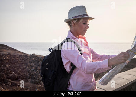 Giovane donna bionda viaggiatore con zaino nero leggere la mappa per scegliere la destinazione successiva. Costa e l'oceano in background. Esterno il concetto di viaggio Foto Stock