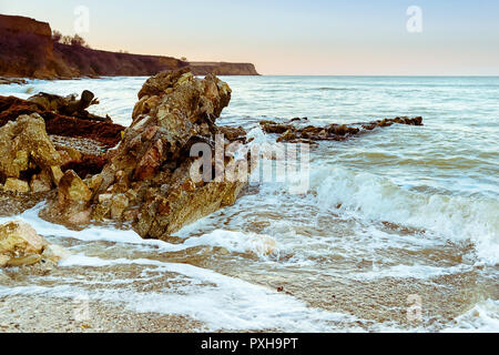 L'onda avvolge la riva, le onde si infrangono su una grande pietra, un luminoso cape in background, foschia al tramonto Foto Stock