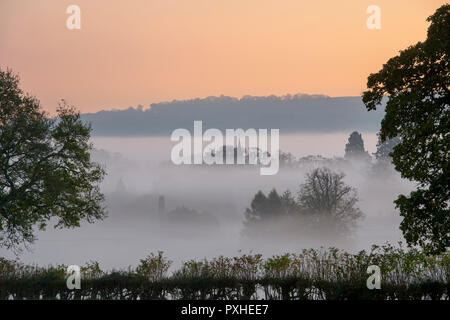 La nebbia e la nebbia a sunrise ribaltamento la campagna intorno al villaggio Costwold di macellazione inferiore in autunno. Cotswolds, Gloucestershire, Inghilterra Foto Stock