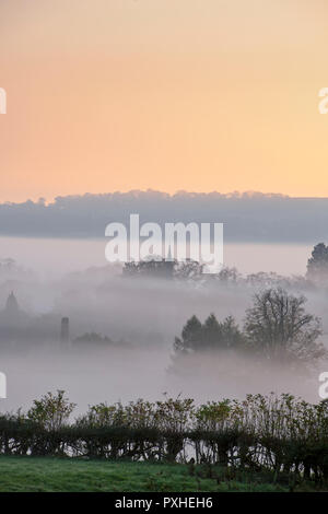 La nebbia e la nebbia a sunrise ribaltamento la campagna intorno al villaggio Costwold di macellazione inferiore in autunno. Cotswolds, Gloucestershire, Inghilterra Foto Stock