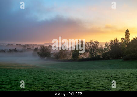 La nebbia e la nebbia a sunrise ribaltamento la campagna intorno al villaggio Costwold di macellazione inferiore in autunno. Cotswolds, Gloucestershire, Inghilterra Foto Stock