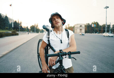 Ritratto di una bionda uomo bianco in città con un classico la bici in spalla, guardando alla videocamera mentre si sta in piedi su una strada in città Foto Stock