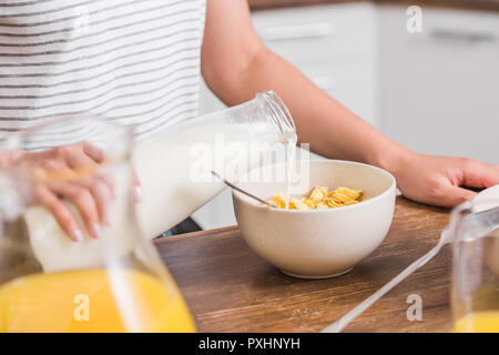 Immagine ritagliata della ragazza versando il latte dalla bottiglia nella piastra con cornflakes durante la colazione nella cucina Foto Stock