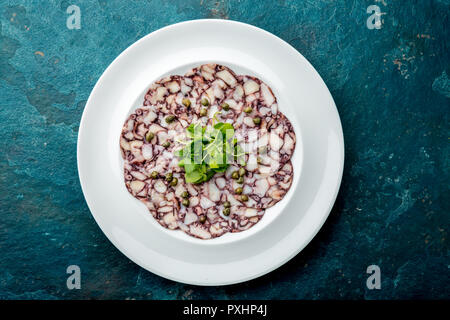 CARPACCIO di polpo. Piatti di pesce crudo fettine di polpo con olio d'oliva, limone e capperi sulla piastra bianca. Vista dall'alto. Sfondo blu Foto Stock