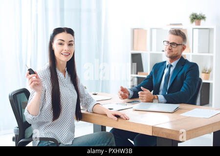 Donna sorridente che mostra auto chiavi in mano mentre è seduto a tavola insieme con agente di assicurazione Foto Stock