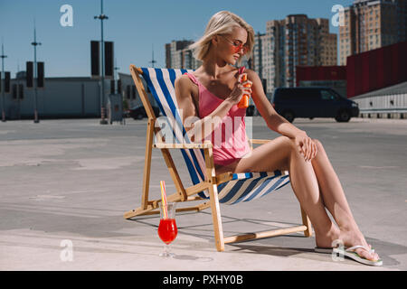 Elegante giovane donna in costume da bagno rosa seduto sulla sedia a sdraio e l'applicazione di crema di protezione solare su parcheggio Foto Stock