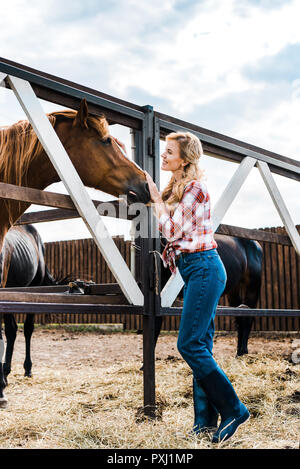 Vista laterale di attraenti sorridente agricoltore palming cavallo in stabile Foto Stock