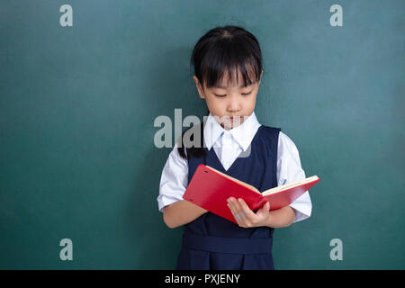 Cinese asiatici bambina in uniforme la lettura del libro verde contro la lavagna in aula Foto Stock