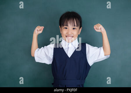 Cinese asiatici bambina in uniforme in piedi con le braccia in alto contro la lavagna verde Foto Stock