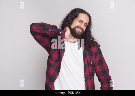 Ritratto di triste uomo bello con barba nera e lunghi capelli ricci in stile casual, a scacchi maglietta rossa in piedi e tenendo la sua dolorosa del collo con clos Foto Stock