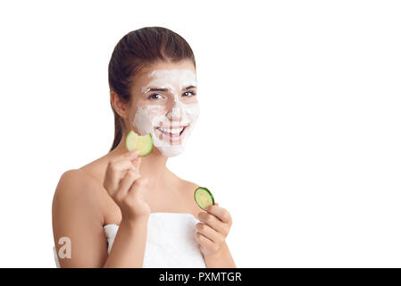 Sorridente attraente giovane donna in un asciugamano che indossa una maschera per il viso trattamento tenendo su fette di cetriolo crudo per i suoi occhi in un centro termale e di bellezza concetto iso Foto Stock