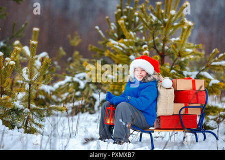 Carino piccolo ragazzo in Santa hat porta una slitta di legno con doni in boschi innevati Foto Stock