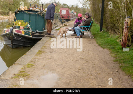 Persone sedute in sedie e bere il tè al fianco di un narrowboat ormeggiato sul canale. Un anziano uomo in piedi il suo cane ha un arresto di una sessione di conversazione testuale. Foxton. Foto Stock