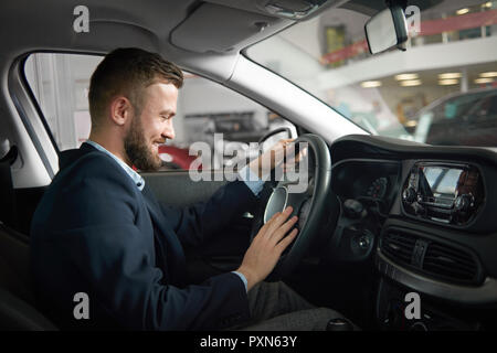 Felice client maschio del centro auto seduto in cabina e di sperimentazione di nuove automobili. Uomo bello indossare in blu scuro tuta tenendo le mani sul volante e lo sta guardando. Foto Stock