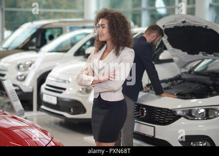 Arricciato donna felice a piedi in auto store, quando il concessionario cerca all'interno di automobile. Client di donna con capelli ricci scegliendo e osservando le vetture per l'acquisto. Manager che lavora in background. Foto Stock