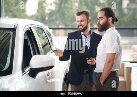 Sorridente manager indossa in dark blue Jacket lavora con il cliente nel centro di auto. Brutale uomo barbuto che indossa in maglia bianca scegliendo e guardando all'automobile. Il concessionario tenendo la penna e la cartella. Foto Stock