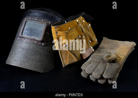 Una vecchia maschera una tuta protettiva per una saldatrice. La salute e la sicurezza sul lavoro abbigliamento per lavoratori di produzione su un tavolo di officina. Sfondo nero. Foto Stock