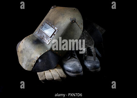 Una vecchia maschera una tuta protettiva per una saldatrice. La salute e la sicurezza sul lavoro abbigliamento per lavoratori di produzione su un tavolo di officina. Sfondo nero. Foto Stock