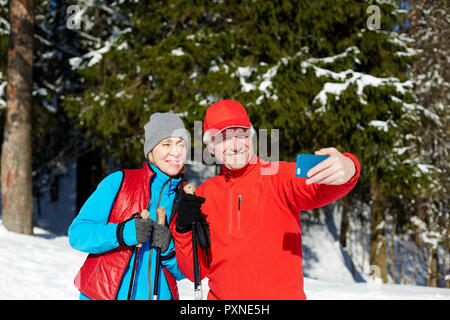 Felice di anziani in inverno activewear rendendo selfie sullo smartphone durante la formazione dello sci nella foresta Foto Stock