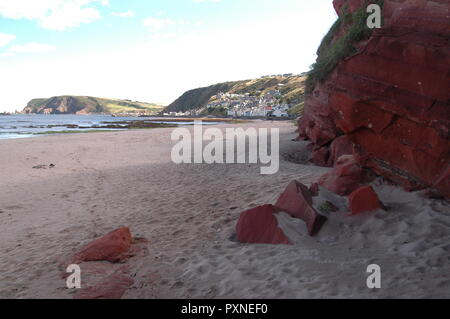 Colore rosso scogliera di arenaria e rocce sulla spiaggia Gardenstown con il villaggio visibile in background Foto Stock