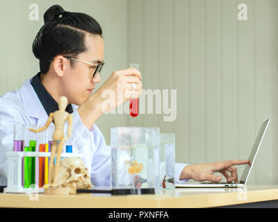 Scienziato asiatico indossando eyeglassess tubo di contenimento contengono liquido rosso Siam witht lotta contro la pesca nel serbatoio durante la registrazione del risultato di test in notebook Foto Stock