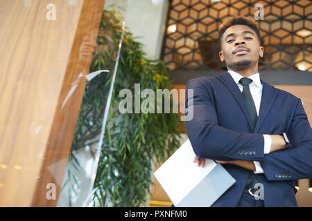 Ritratto di fiducioso giovane imprenditore africani in abbigliamento formale in piedi con documenti presso un ufficio moderno Foto Stock