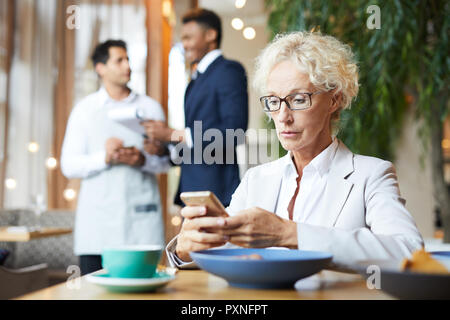 Grave imprenditrice matura in occhiali, utilizzando il suo smartphone mentre si attende per il caffè cameriere o partner presso il ristorante Foto Stock