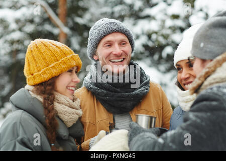 Allegro eccitato giovani escursionisti in cappelli in piedi in cerchio e ridere mentre chiacchierare insieme e di bere il tè caldo durante le escursioni nella foresta di inverno Foto Stock