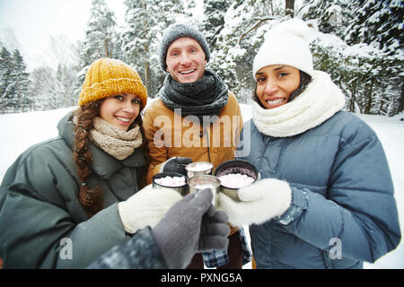 Felice giovani multi-etnico amici in caldi cappotti tintinnio thermos tazze mentre il riscaldamento da bere una bevanda con marshmallows in inverno la foresta, essi cercando Foto Stock