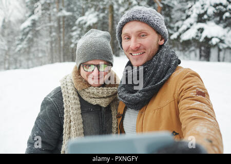 Positivo bel giovane gli uomini in cappelli e sciarpe in piedi nella foresta di inverno e rendendo Self-portrait sullo smartphone Foto Stock