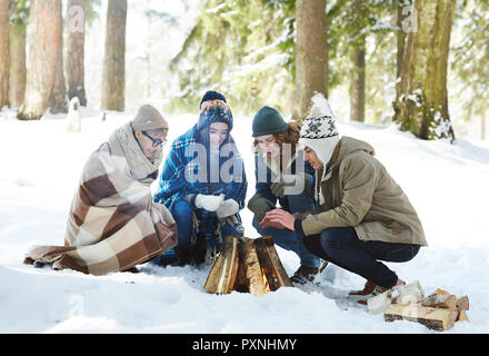 A piena lunghezza Ritratto di quattro giovani camping in inverno forest seduti in cerchio intorno il fuoco Foto Stock