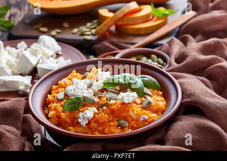 Close-up di crema di zucca butternut e purea di feta con riso e semi di zucca e di foglie di basilico fresco su una piastra di terracotta con gli ingredienti su di un legno Foto Stock