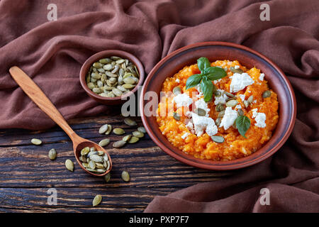 Crema di zucca butternut e purea di riso con formaggio feta, semi di zucca e di foglie di basilico fresco in un vaso di argilla con ingredienti su un rustico in legno tabl Foto Stock