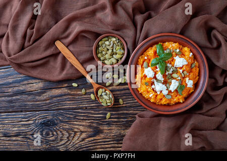 Crema di zucca butternut e purea di riso con formaggio feta, semi di zucca e di foglie di basilico fresco in un vaso di argilla su una tavola di legno tavolo rustico, vista da abo Foto Stock