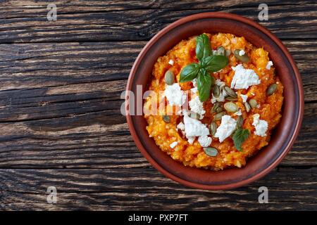 Crema di zucca butternut e purea di riso con formaggio feta, semi di zucca e di foglie di basilico fresco in un vaso di argilla su una tavola di legno tavolo rustico, vista da abo Foto Stock