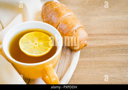 La mattina la colazione con croissant freschi e la tazza di tè con limone su una tavola di legno di colore arancione. Foto Stock