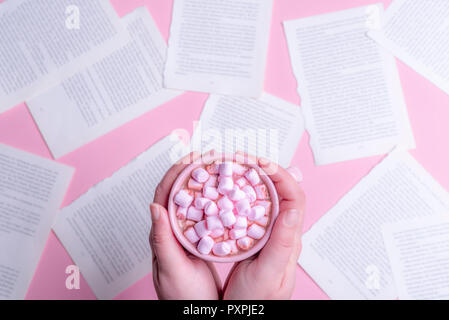 Donna che mantiene, con entrambe le mani, una rosa di tazza di cioccolata calda con mini rosa marshmallows, al di sopra di un tavolo pieno di pagine di un libro. Foto Stock